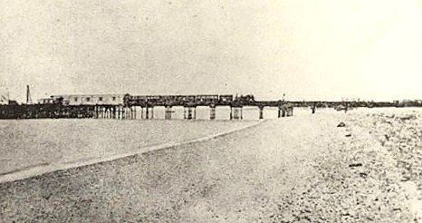 A black and white image shows a shoreline with a railway pier extending to the left into the sea. On the pier a steam train can be seen.