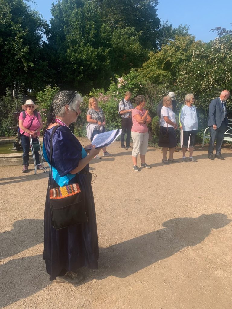 In the foreground is a lady dressed as Queen Victoria. She is wearing and amplifying headset. She is talking to a group of walkers in the midground. The setting is an ornamental garden.aa