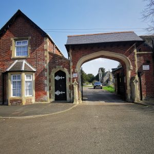 To the left is a brick lodge on two floors with bay window on the ground floor. The lodge is connected to the right with an arched pedestrian door. This door forms part of the main gate which is a brick structure with a flatten arch to the top and above that a tiled roof. Through the archway a car and a Gothic Chapel of Rest can be seen