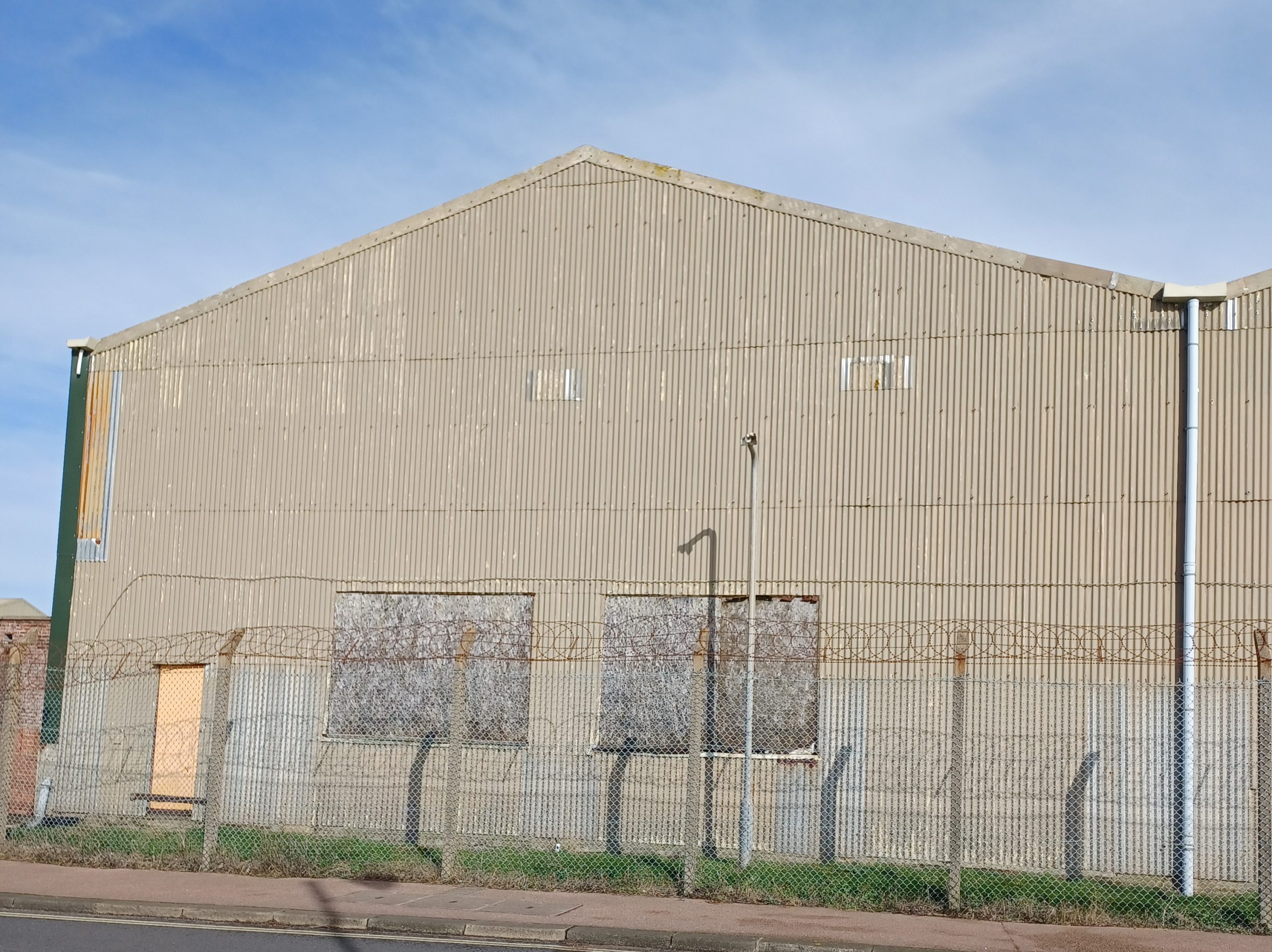 A large hangar made of corrugated steel sits behind a chain-link fence with concrete posts.