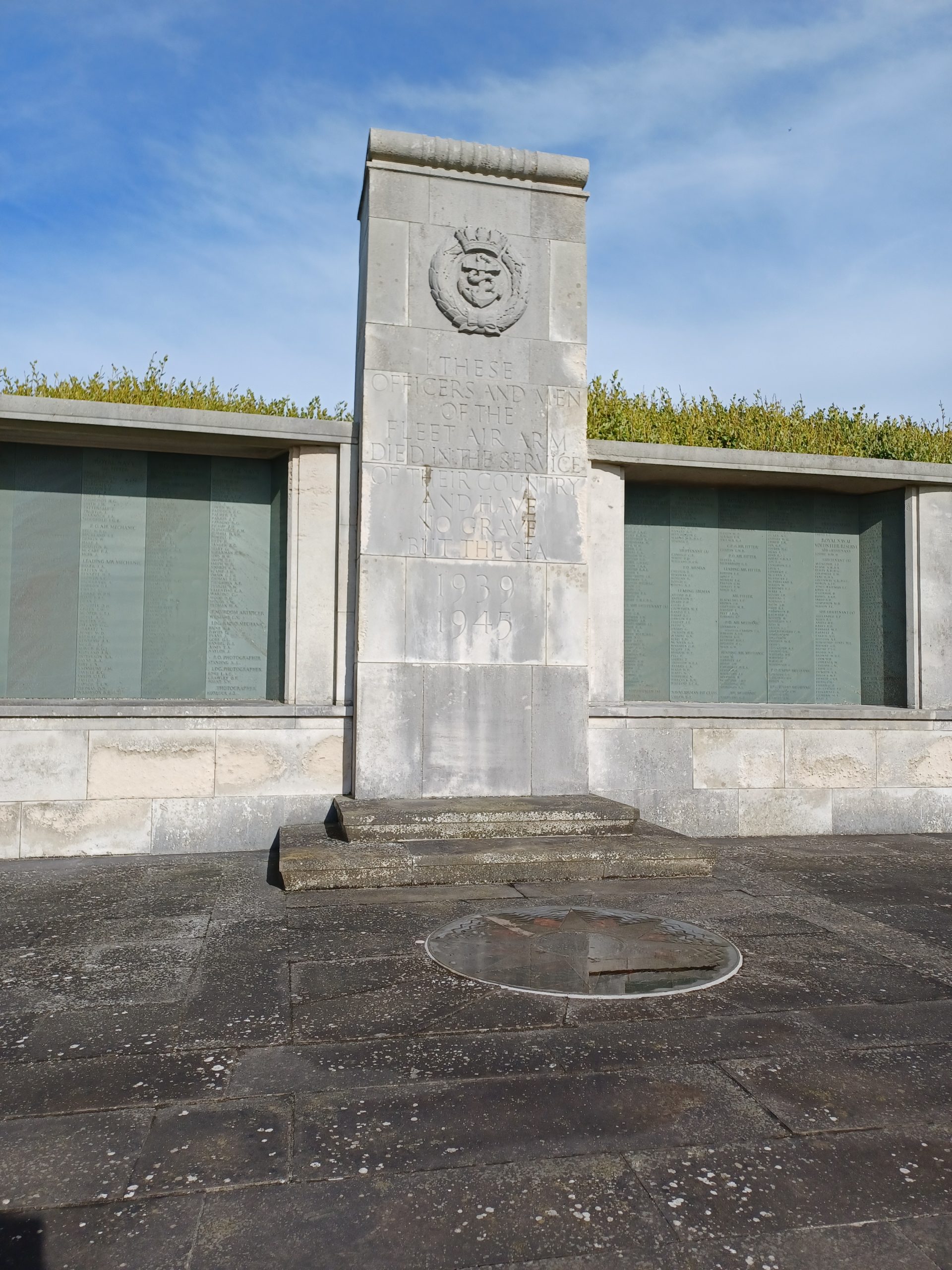 A stone war memorial to the Fleet Air Arm. In the centre is a wide pillar bearing the crest of the FAA. To either side set into the stone are plaques in grey-blue slate bearing the names of those that are remembered