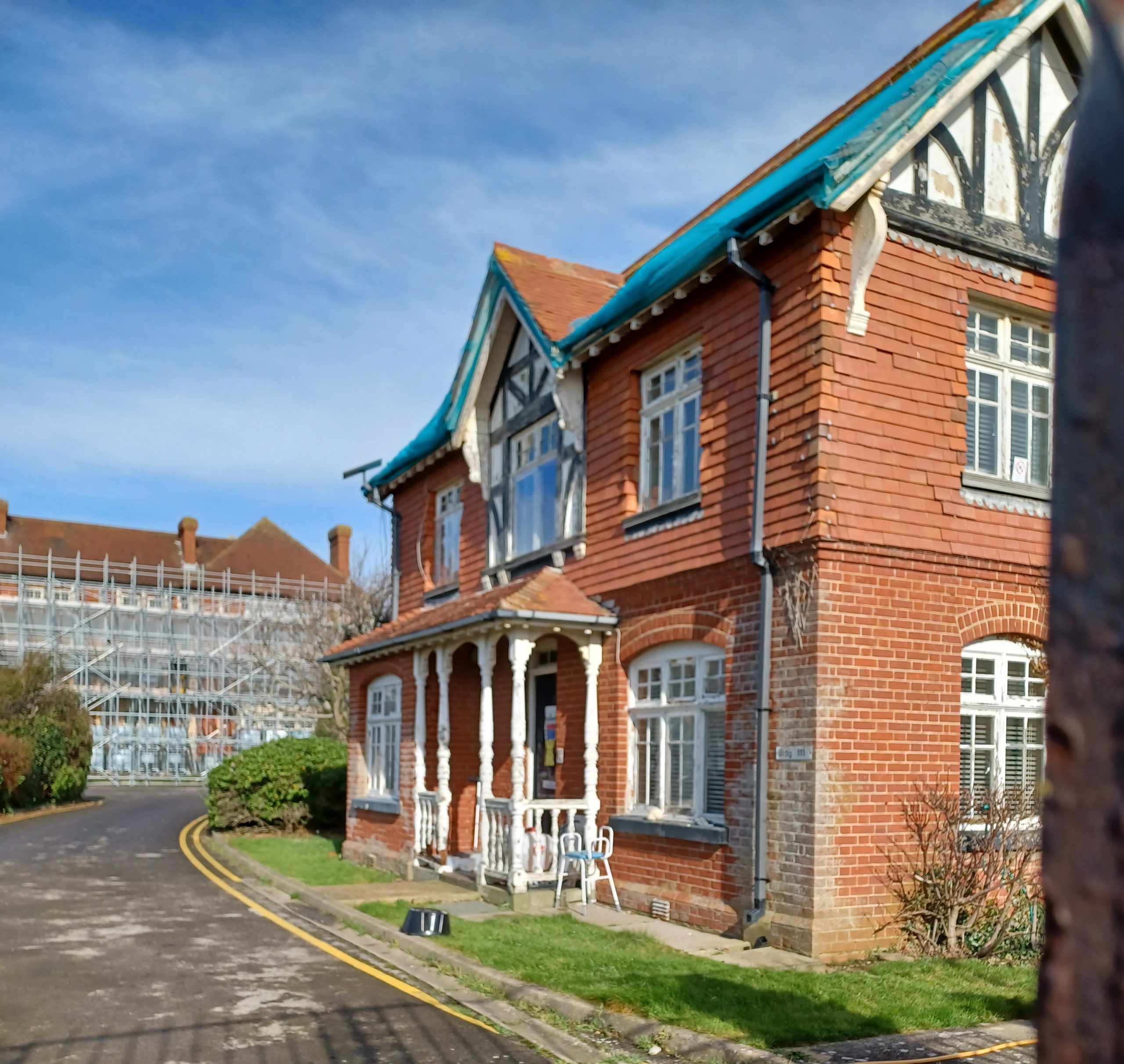 A brick building with Tudor gables. A railed porch is in the centre. In the background can be seen a large scaffolded building.