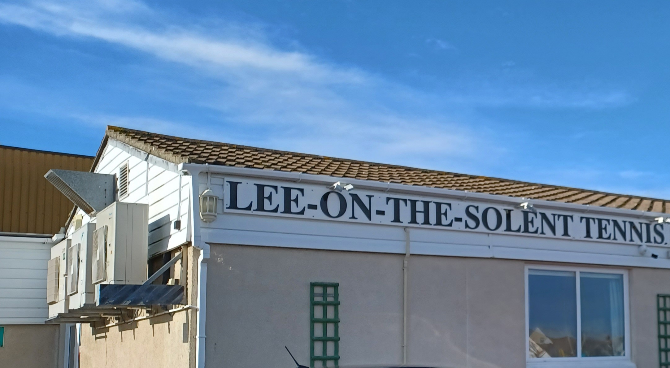 A single storey gambled building. On the end wall to the left are 3 raised air conditioning units. At the top of the Long side of the building a sign reads LEE-ON-THE-SOLENT TENNIS.