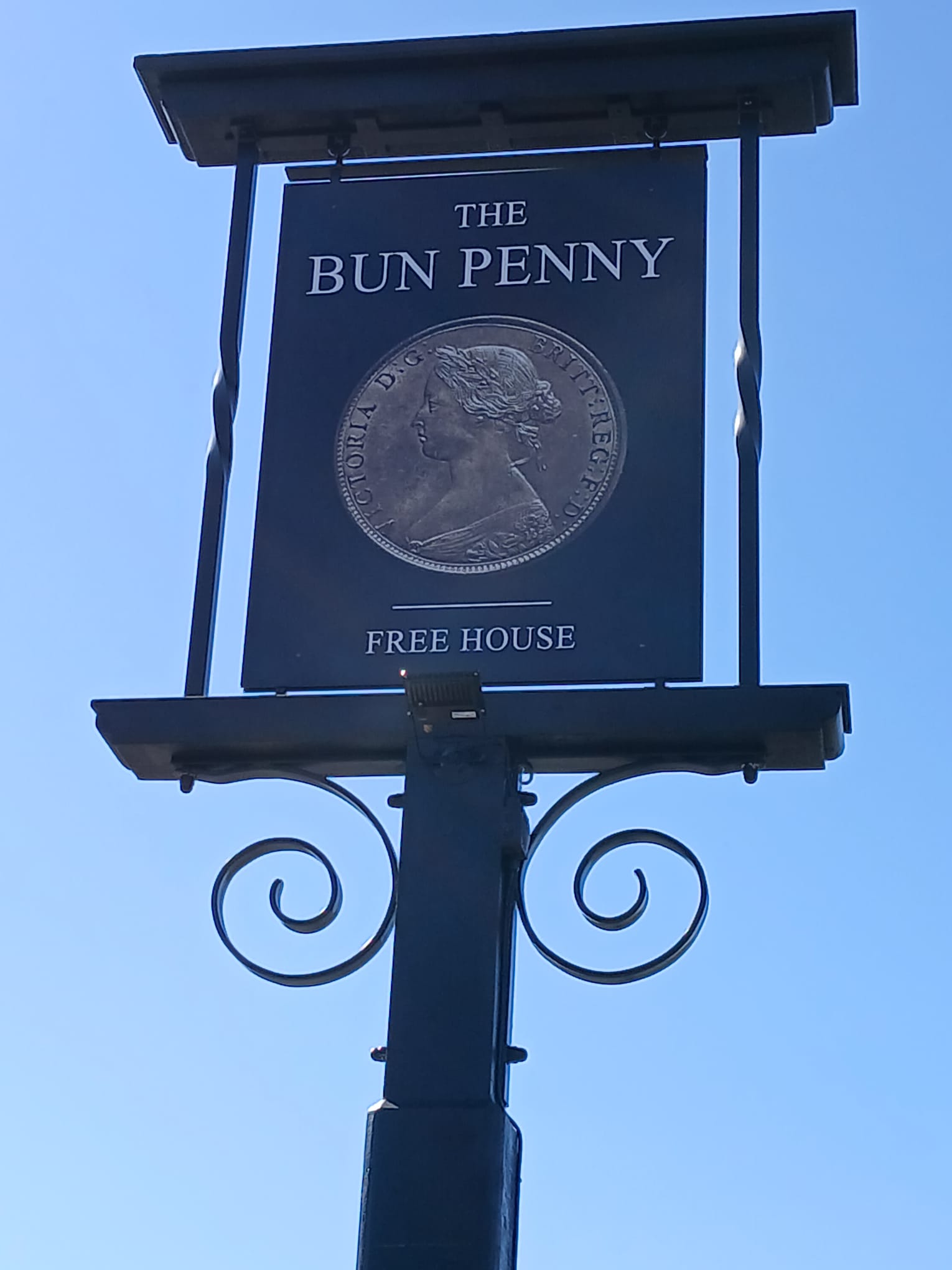 A pub sign hangs from a frame above a large post. The image is of a coin depicting a young Queen Victoria. The sign reads "The Bun Penny, Free House"