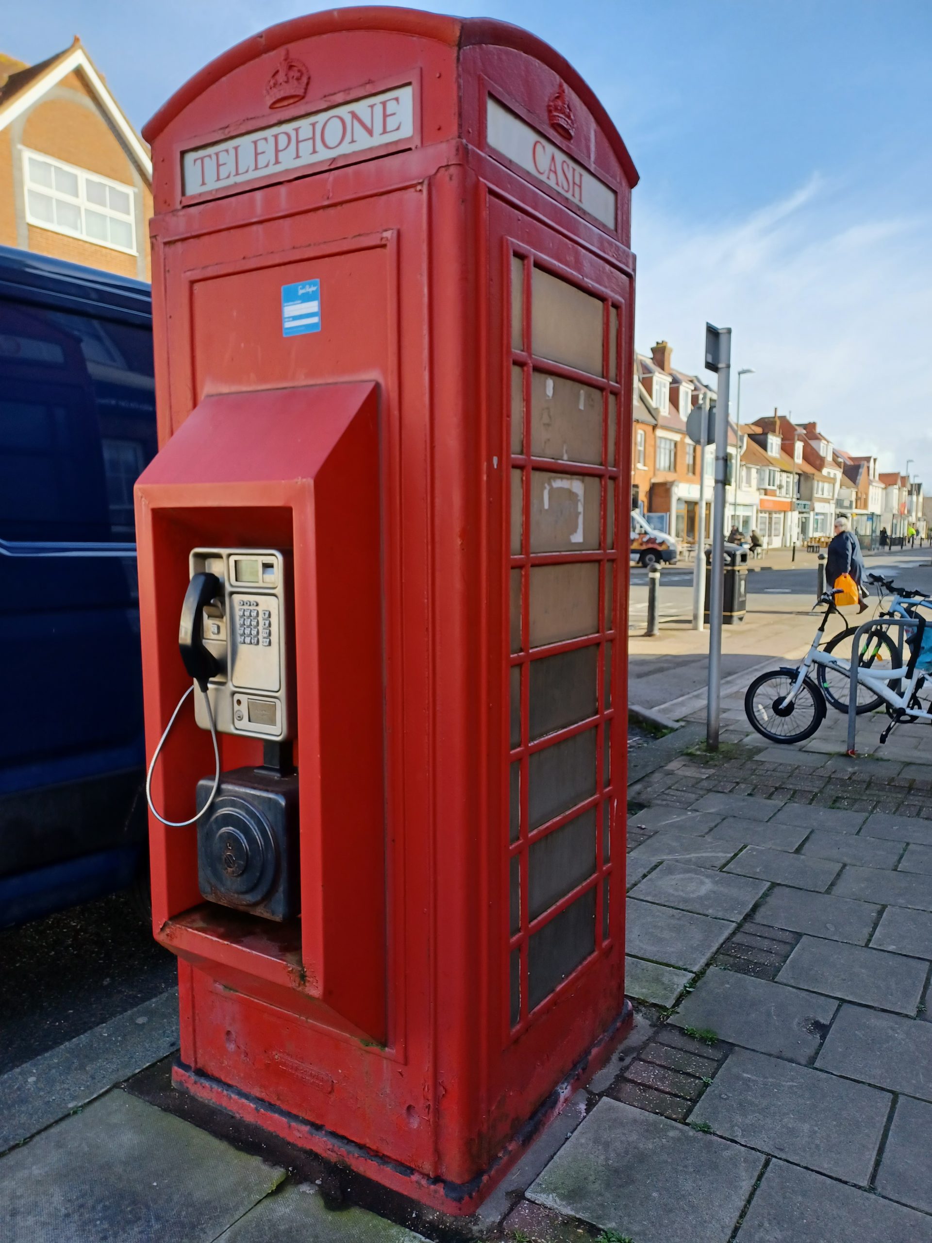 A red phone box in the old style. Converted to a cash machine. A push-button pay phone is mounted on the outside
