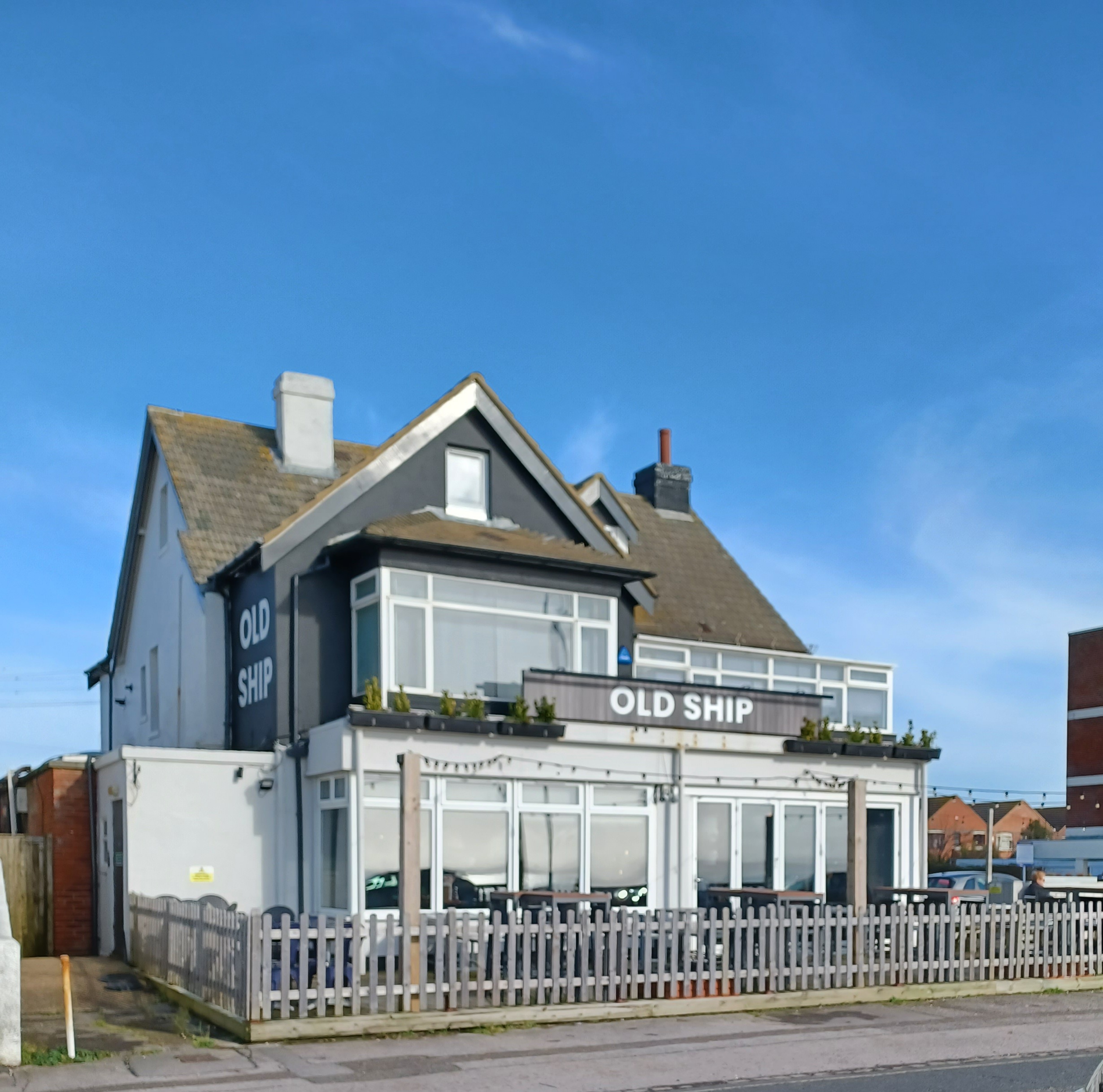 a public house . the sign above the ground floor reads OLD SHIP. In front are tables with benches. A picket fence separates the pub from the pavement.