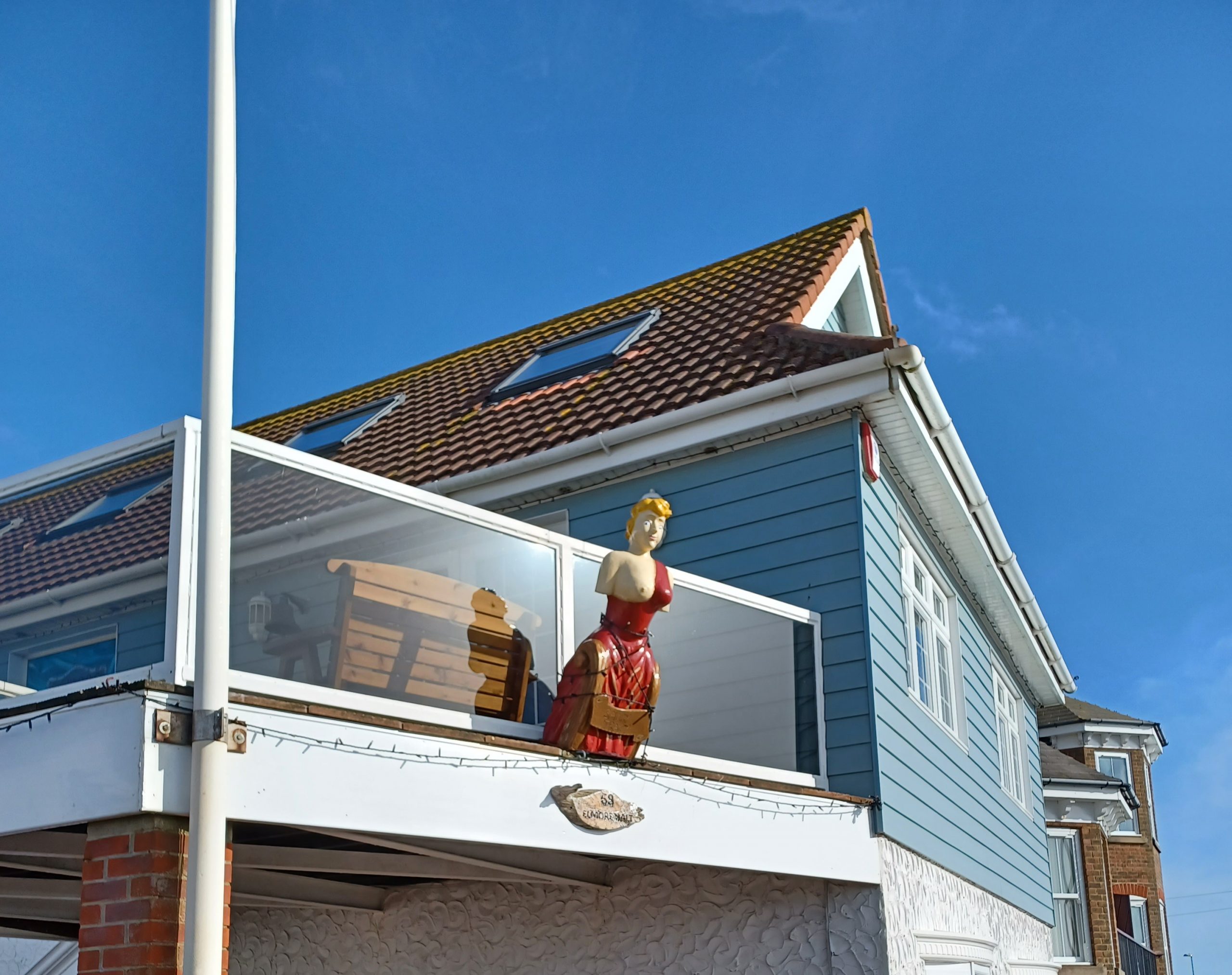 A house with blue weather board on the top storey. At the centre is a ship's figurehead of a woman dressed in red with her right breast uncovered projecting from a glass balcony.