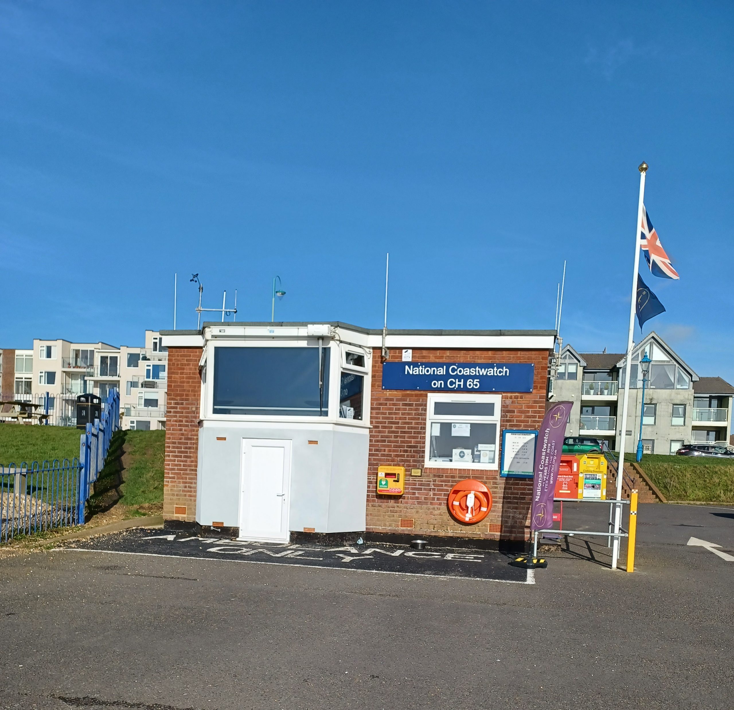 a small brick building with a large bay window. on the wall are an emergency resuscitator and a life belt. to the right of building a flagpole with the Union Flag.