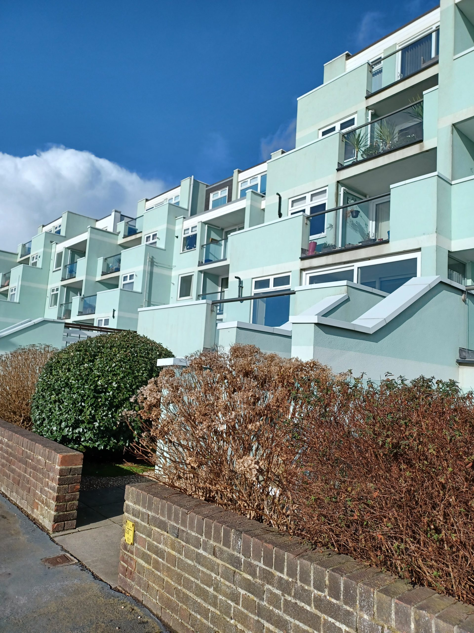 Photo of balconied retirement apartments. The walls are painted pale blue and the balconies have glazed fronts. In the foreground is a low brick wall with a hedge.