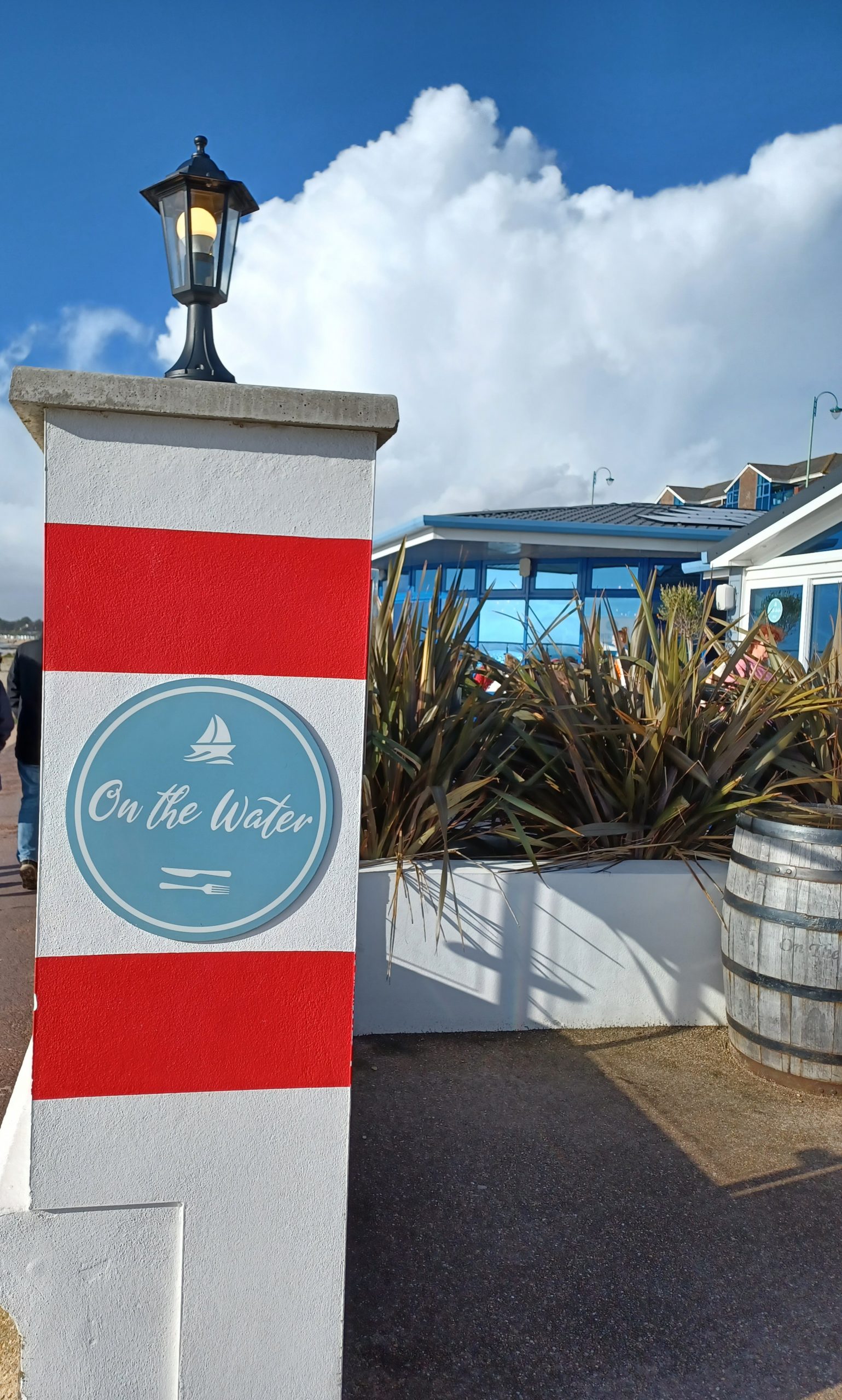on the left is a sign with calligraphic text in white on a circular light blue background with a restaurant building on the right. Text reads "On The Water"